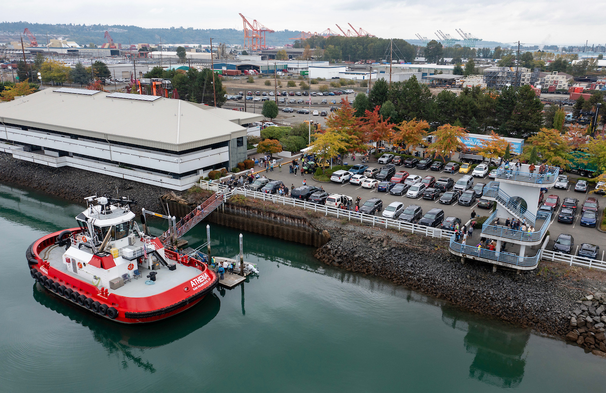 A red tug boat is docked near a parking lot. 