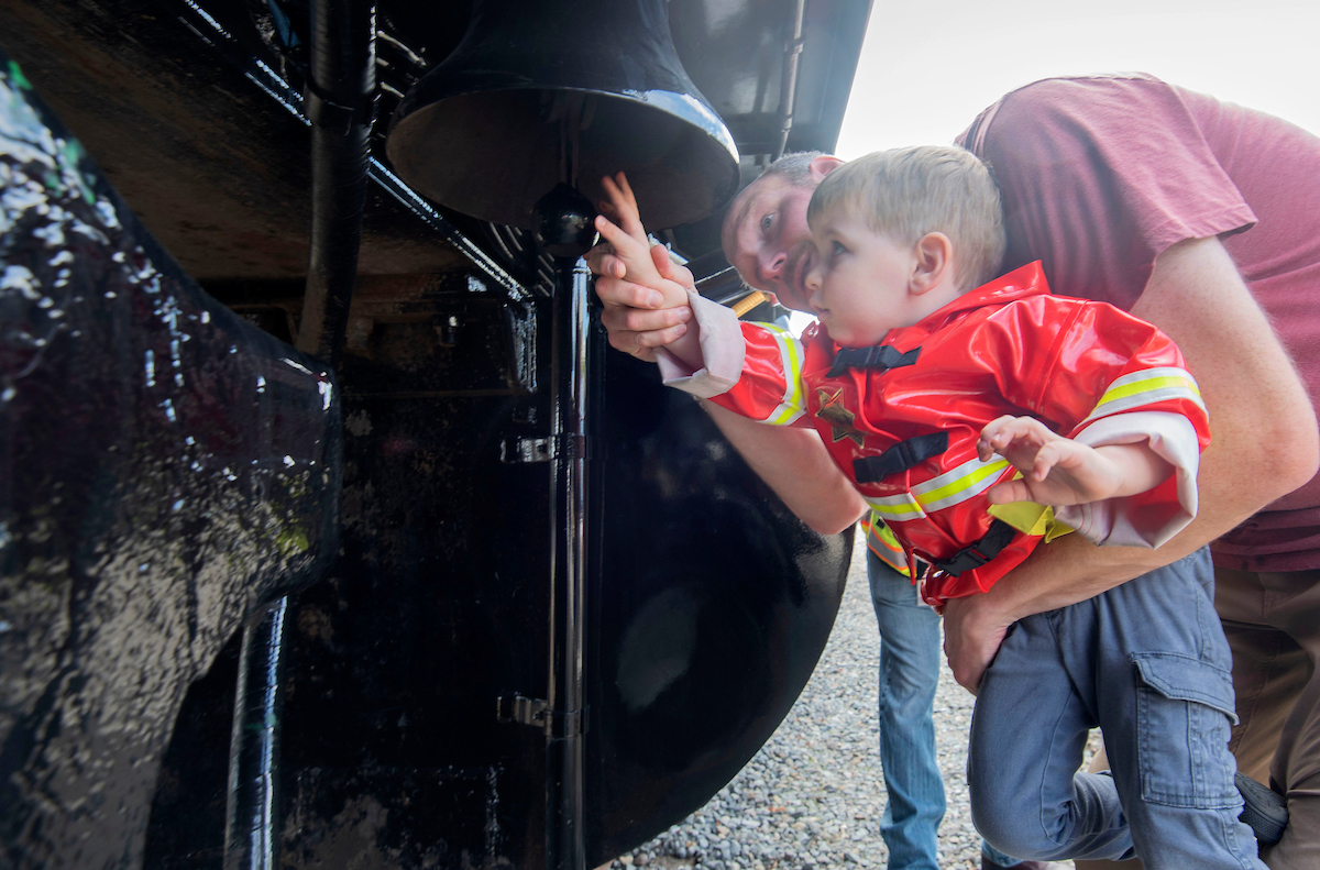 A little boy in a firefighter costume touches parts of a truck while an adult holds them up.
