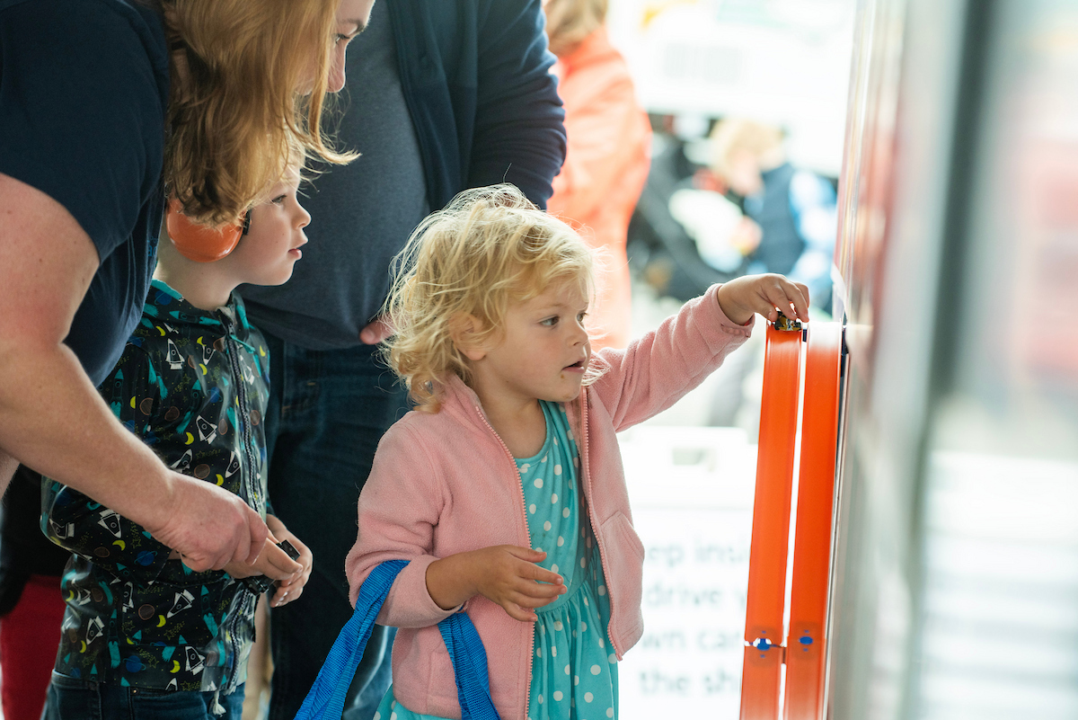 A small girl in a pink coat holds a toy car on an orange track attached to a wall.