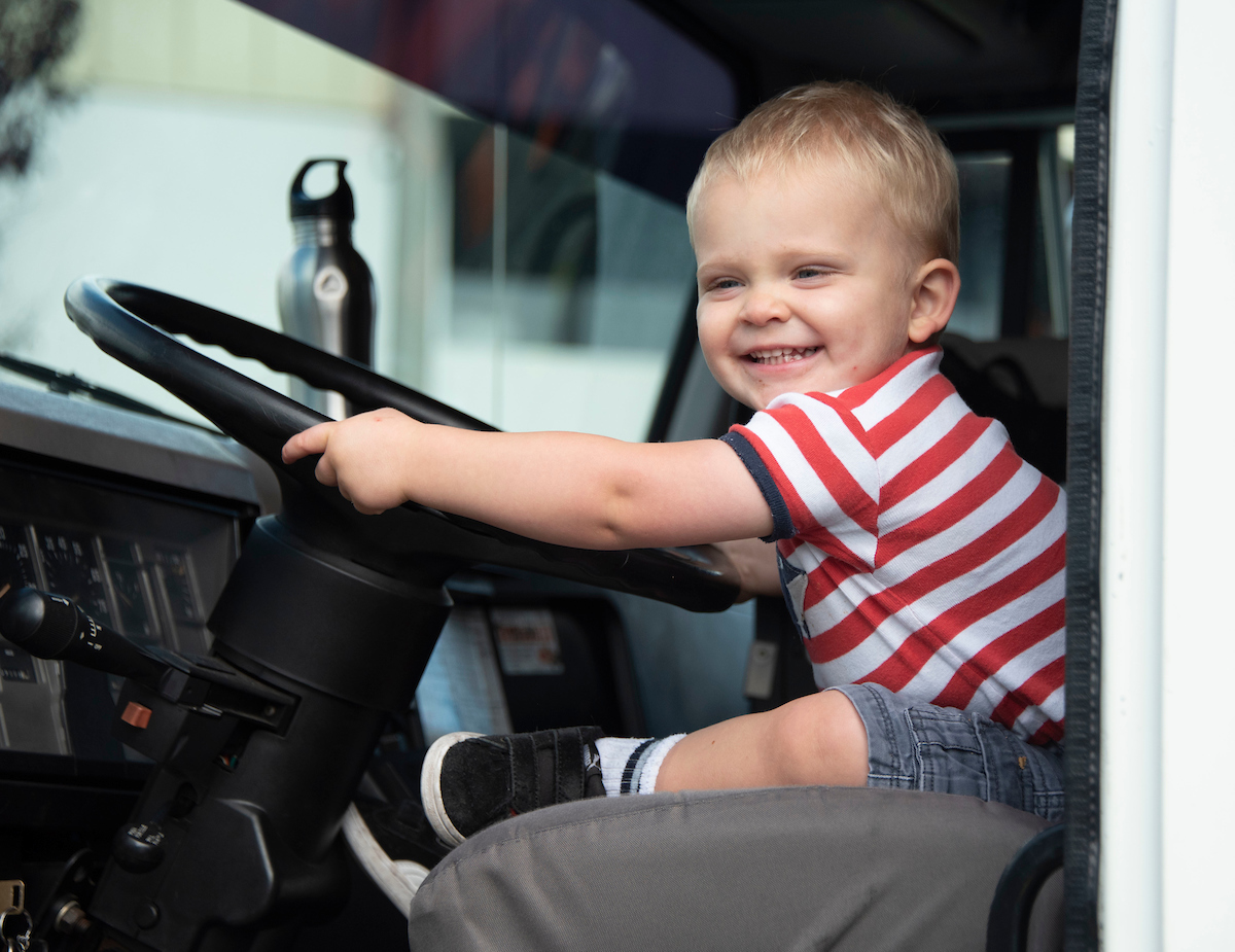 A toddler in a red striped shirt smiles while holding on to the steering wheel of a truck.