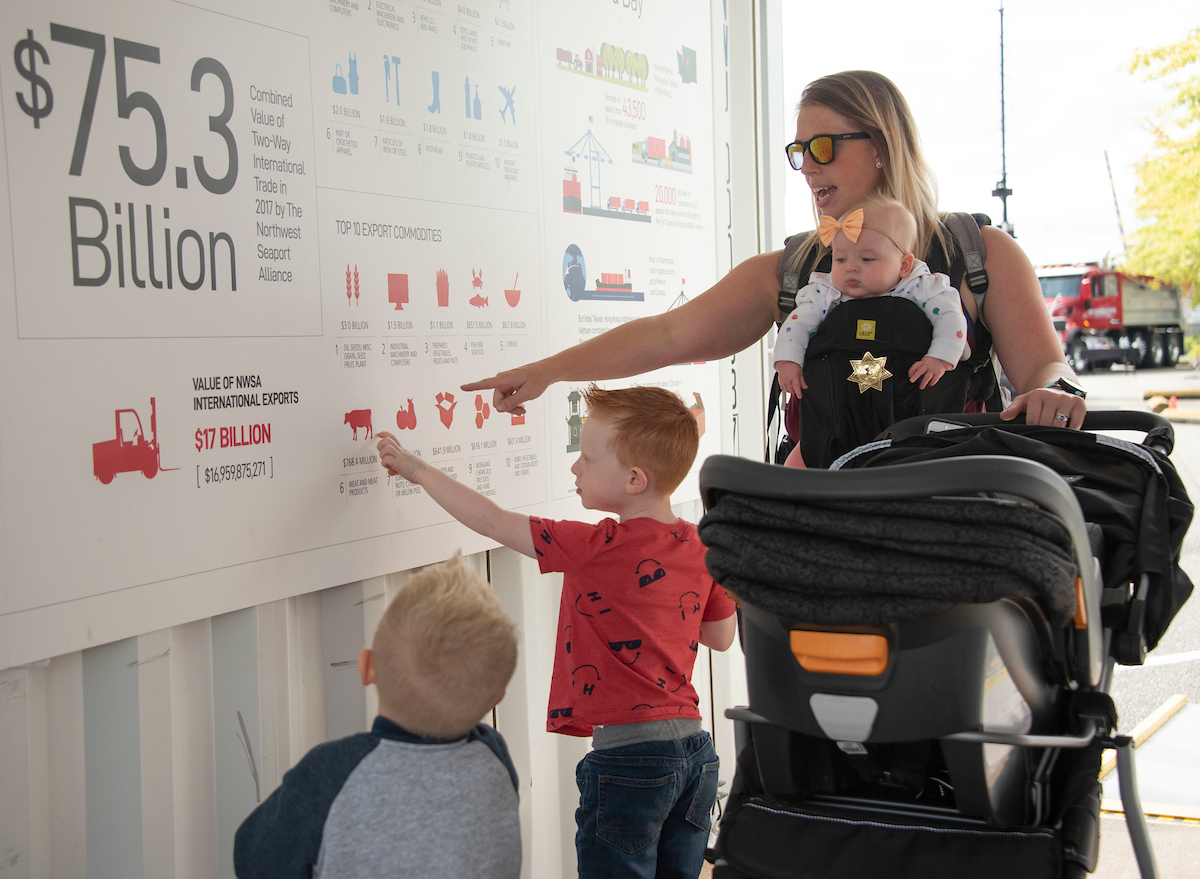A woman holding a baby in a carrier points at a sign for two small boys. 