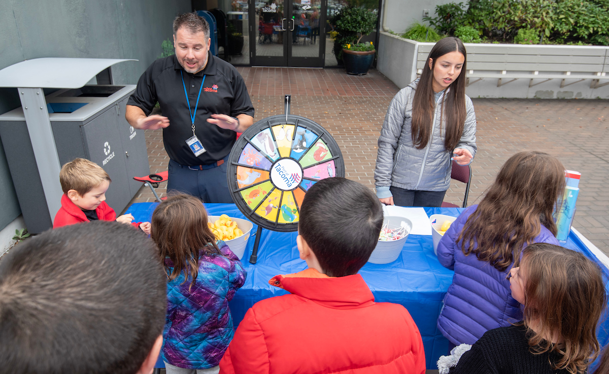 An adult stands at a table with a prize wheel in front of several children.