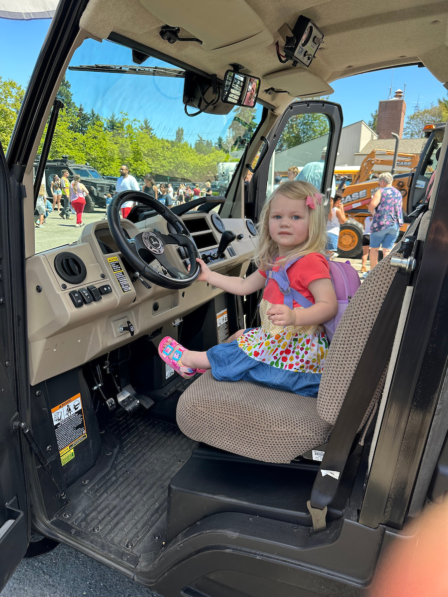 A small blonde girl with a backpack sits in the cab of a truck.