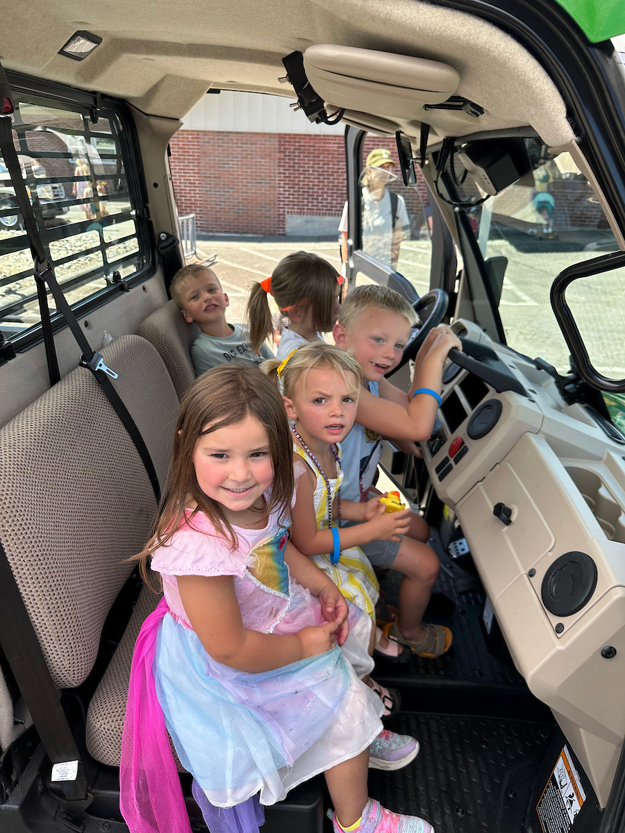 Several small children sit in the cab of a construction vehicle while smiling at the camera. 