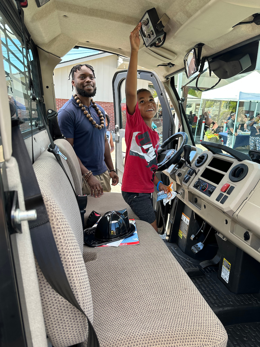 A young boy touches the controls of a construction truck while his father looks on.