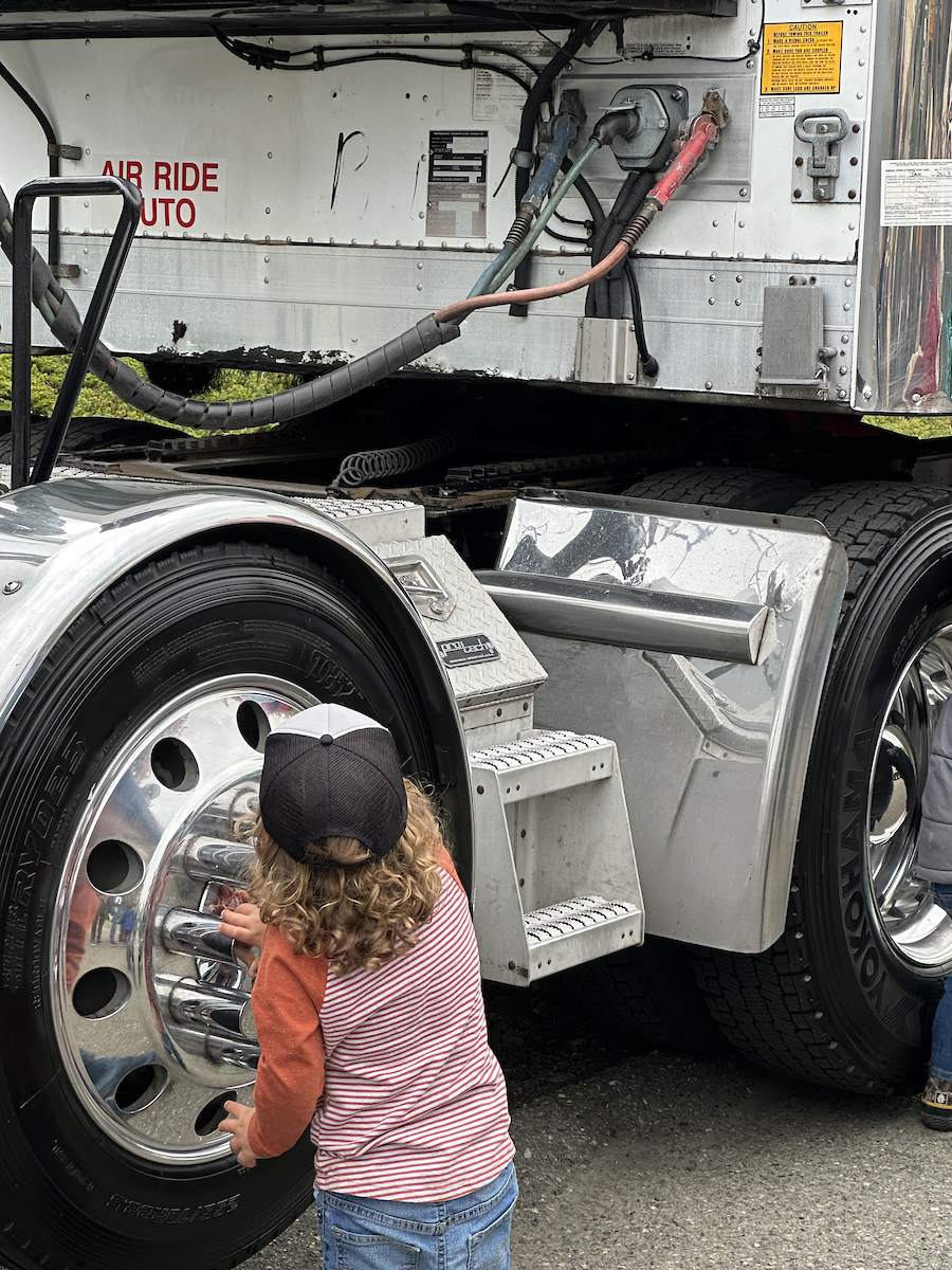 A young child in a hat touches the wheels of a large truck.