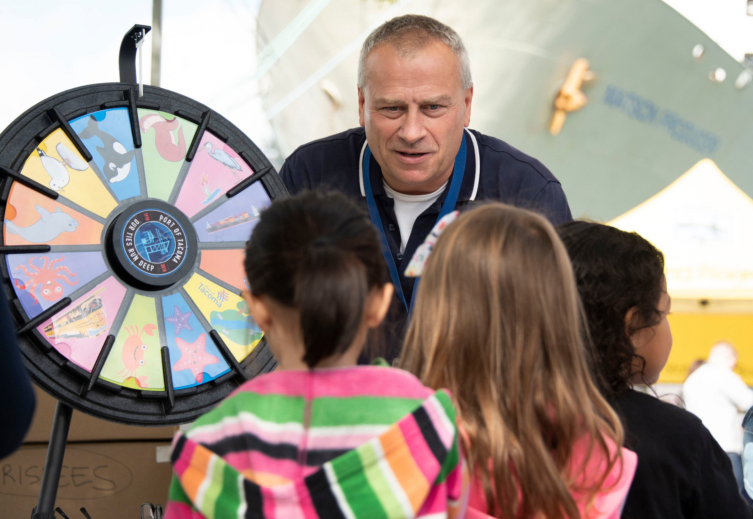 A man leans over a table to ralk to children next to a prize wheel.