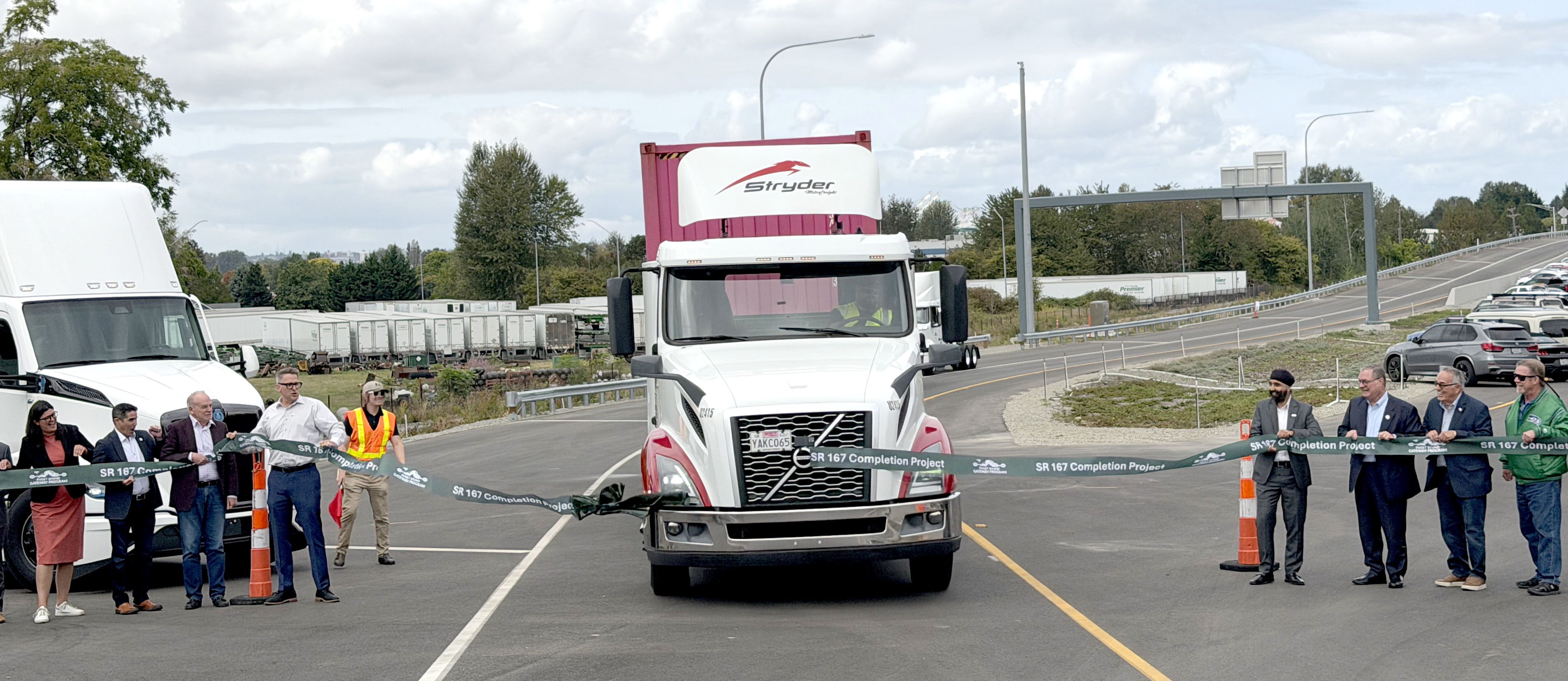 A semi truck drives through a ribbon celebrating the SR167 opening while community partners hold the ribbon.
